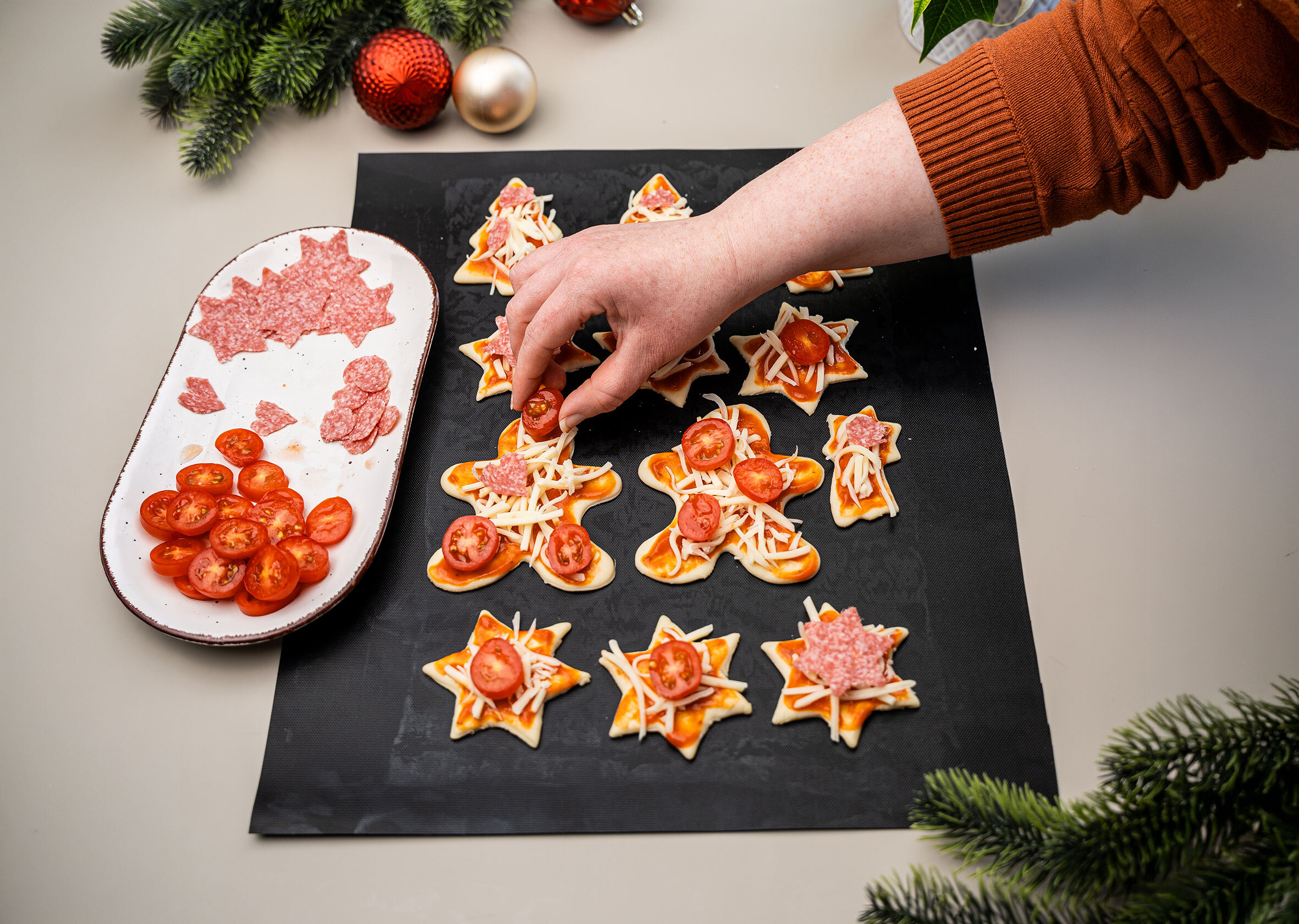 Hände belegen Weihnachtsbaum-, Stern- und Lebkuchenmann-Teiglinge mit Salami und Tomaten.