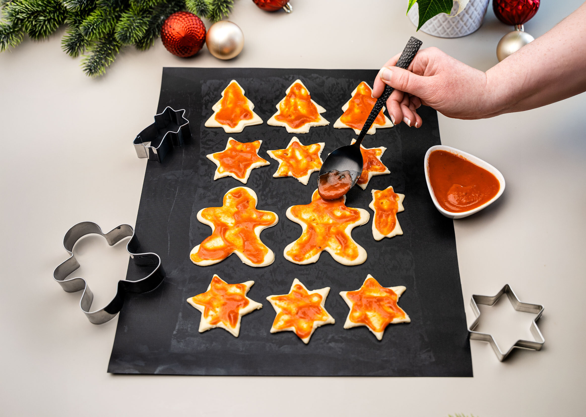 Hände verteilen Tomatensoße auf Weihnachtsbaum-, Stern- und Lebkuchenmann-Teiglinge.