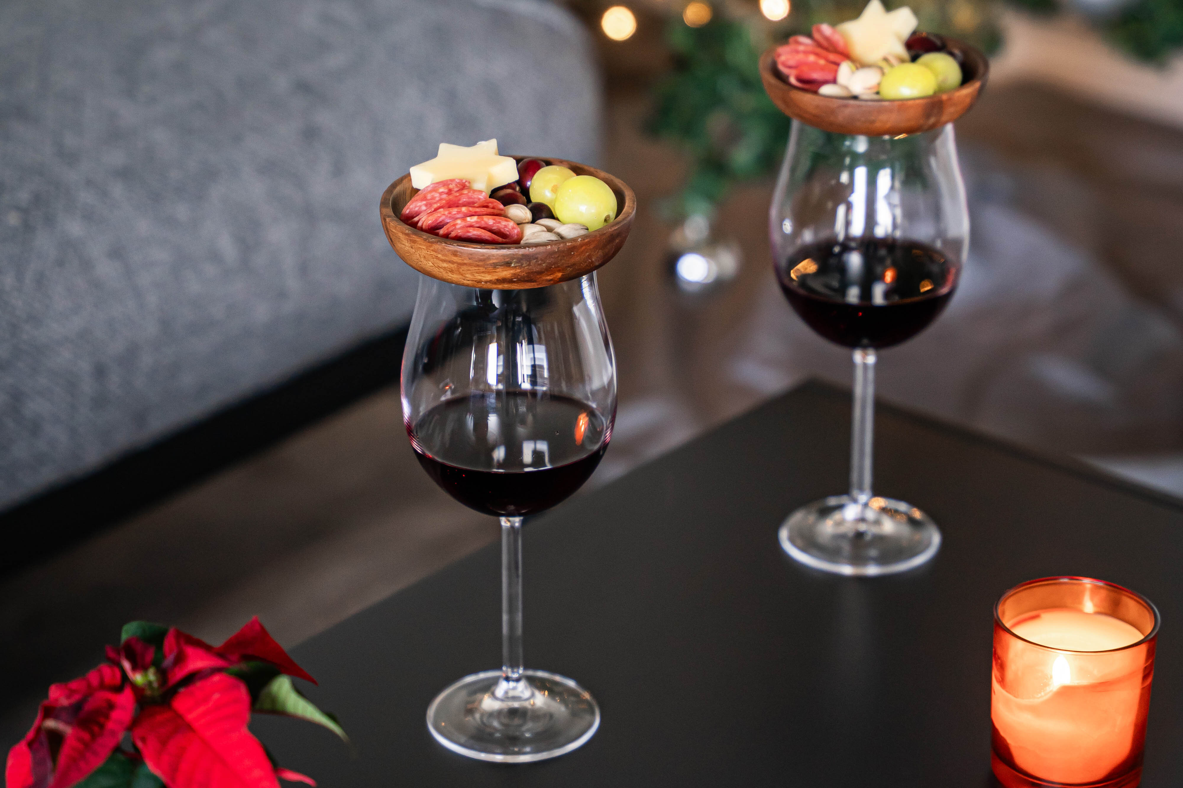 Zwei Weingläser mit rotem Wein, jeweils mit einem Holzbrettchen mit Käse, Trauben und Wurstscheiben darauf, auf einem schwarzen Tisch neben einer brennenden Kerze und einer roten Pflanze.