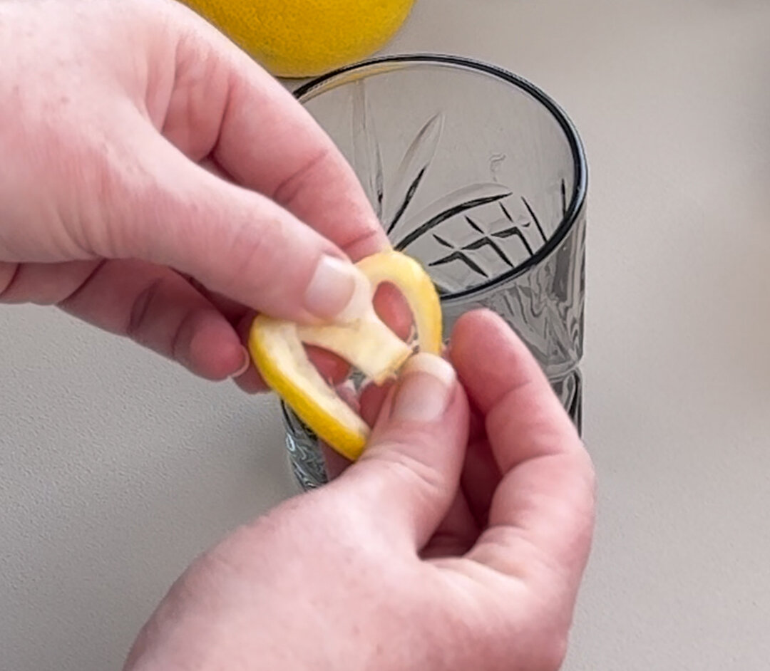 Eine Hand formt eine Zitronenscheibe zu einem Herz und hält sie über ein Glas. Im Hintergrund liegt eine ganze Zitrone und Pflanzenblätter.
