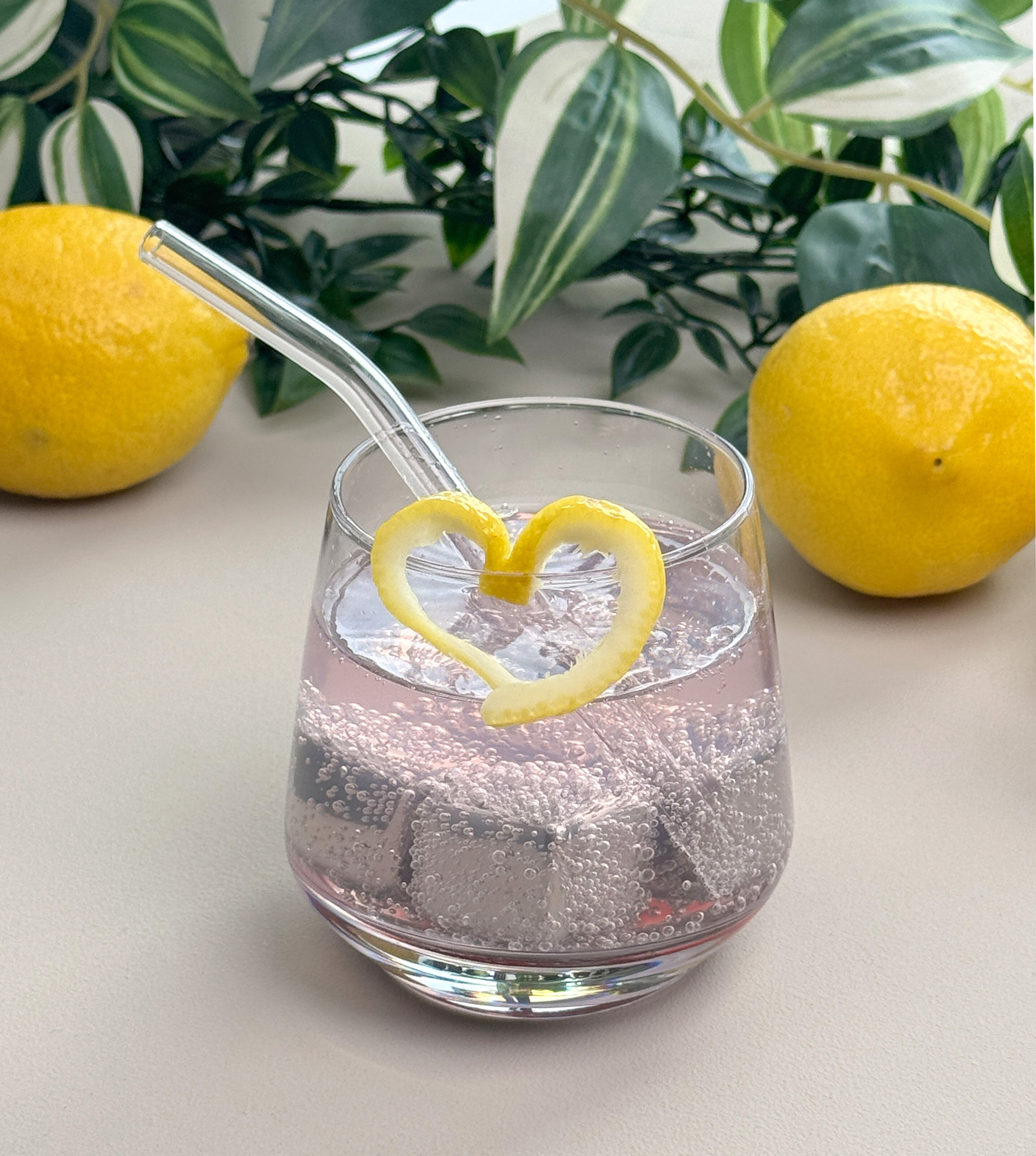 Ein Glas mit sprudelndem Getränk und Eiswürfeln, dekoriert mit einer herzförmigen Zitronenschale. Im Hintergrund liegen zwei Zitronen.