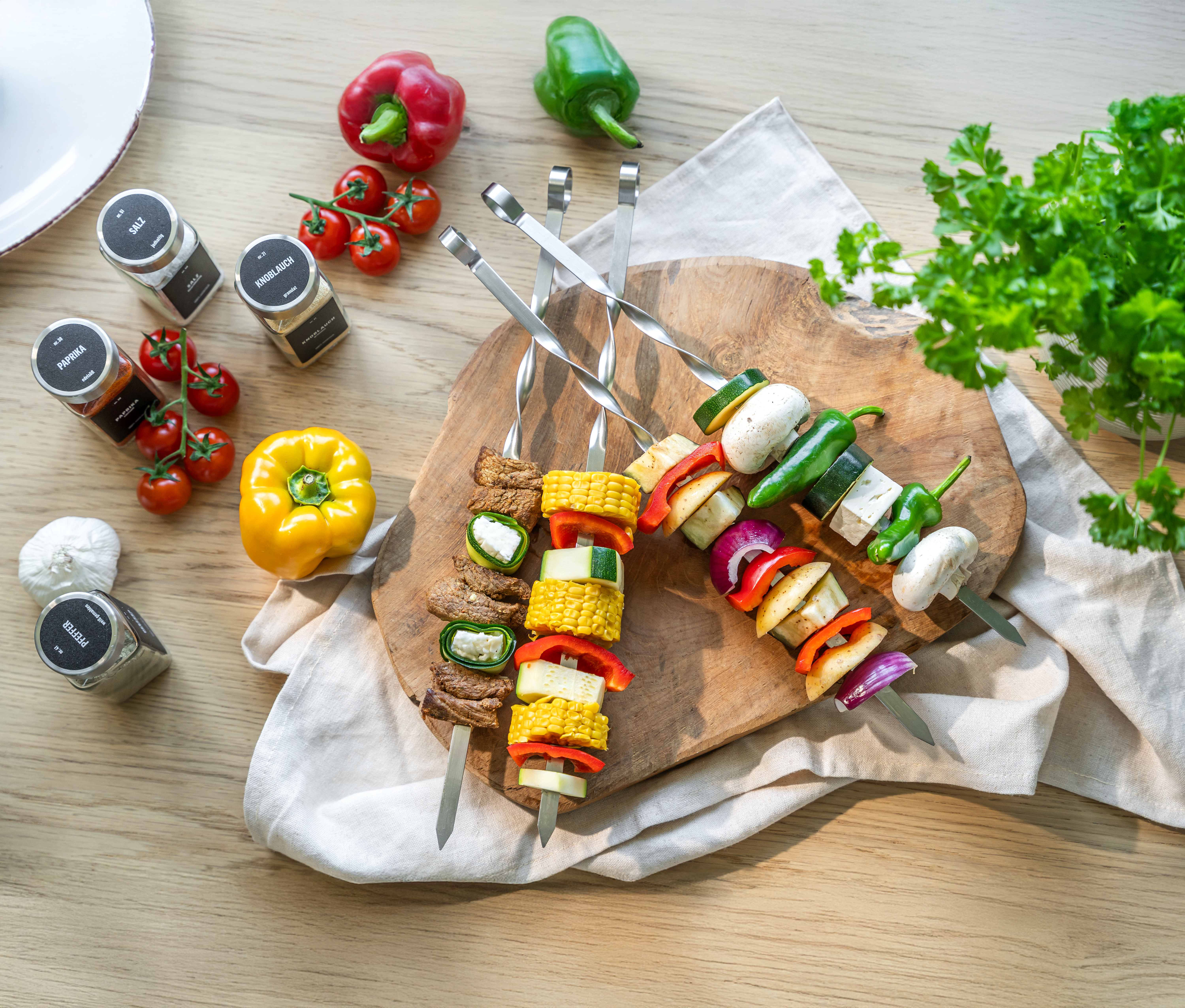 Vier Grillspieße mit Fleisch, Mais, Paprika, Zucchini, Pilzen und Zwiebeln auf einem Holzbrett neben Gewürzgläsern, Paprika, Kirschtomaten und frischer Petersilie