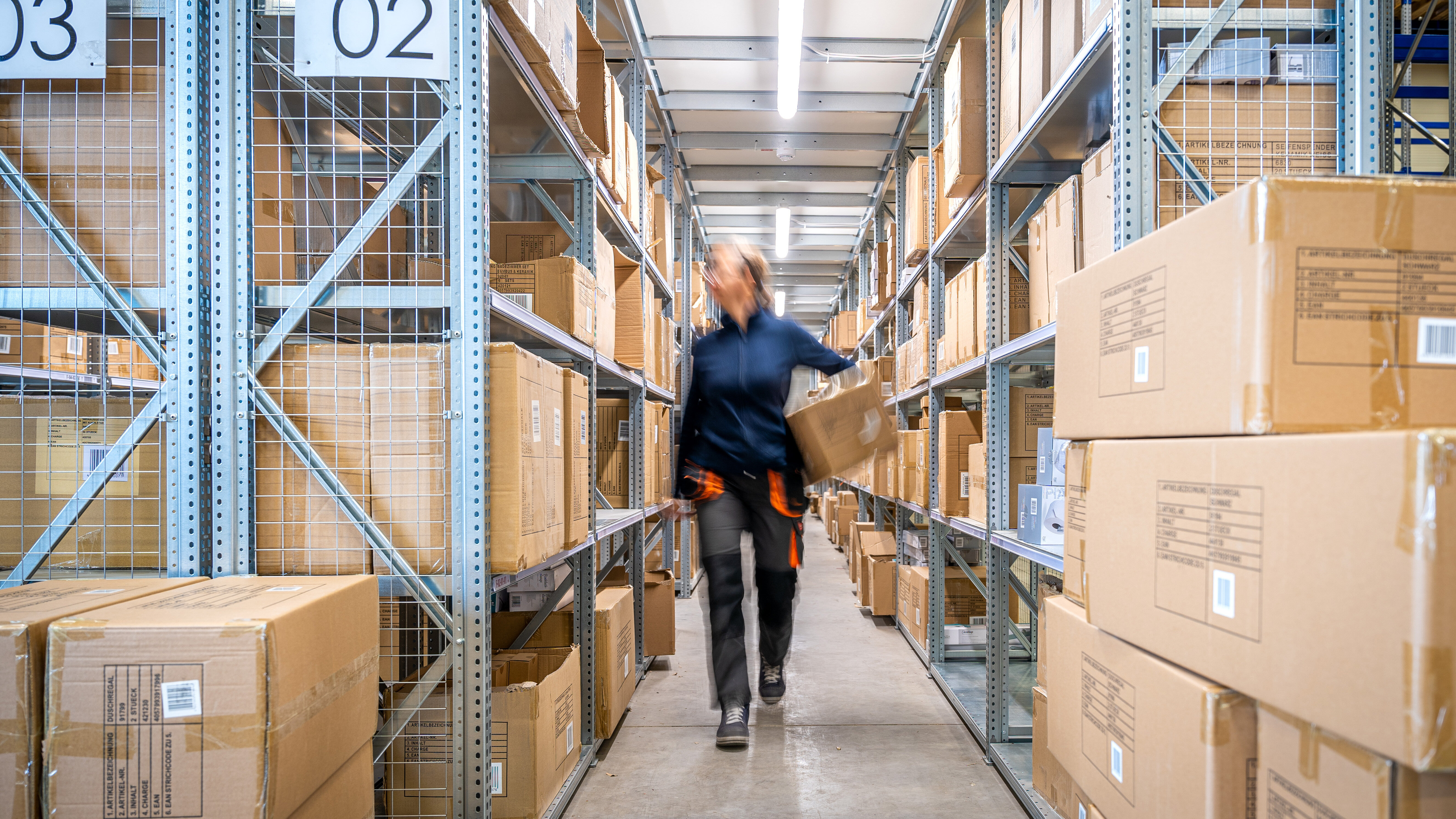 Ein Lagerarbeiter trägt einen Karton durch einen Gang zwischen hohen Regalen, die mit Kartons gefüllt sind. Die Regale sind aus Metall und die Kartons sind braun.