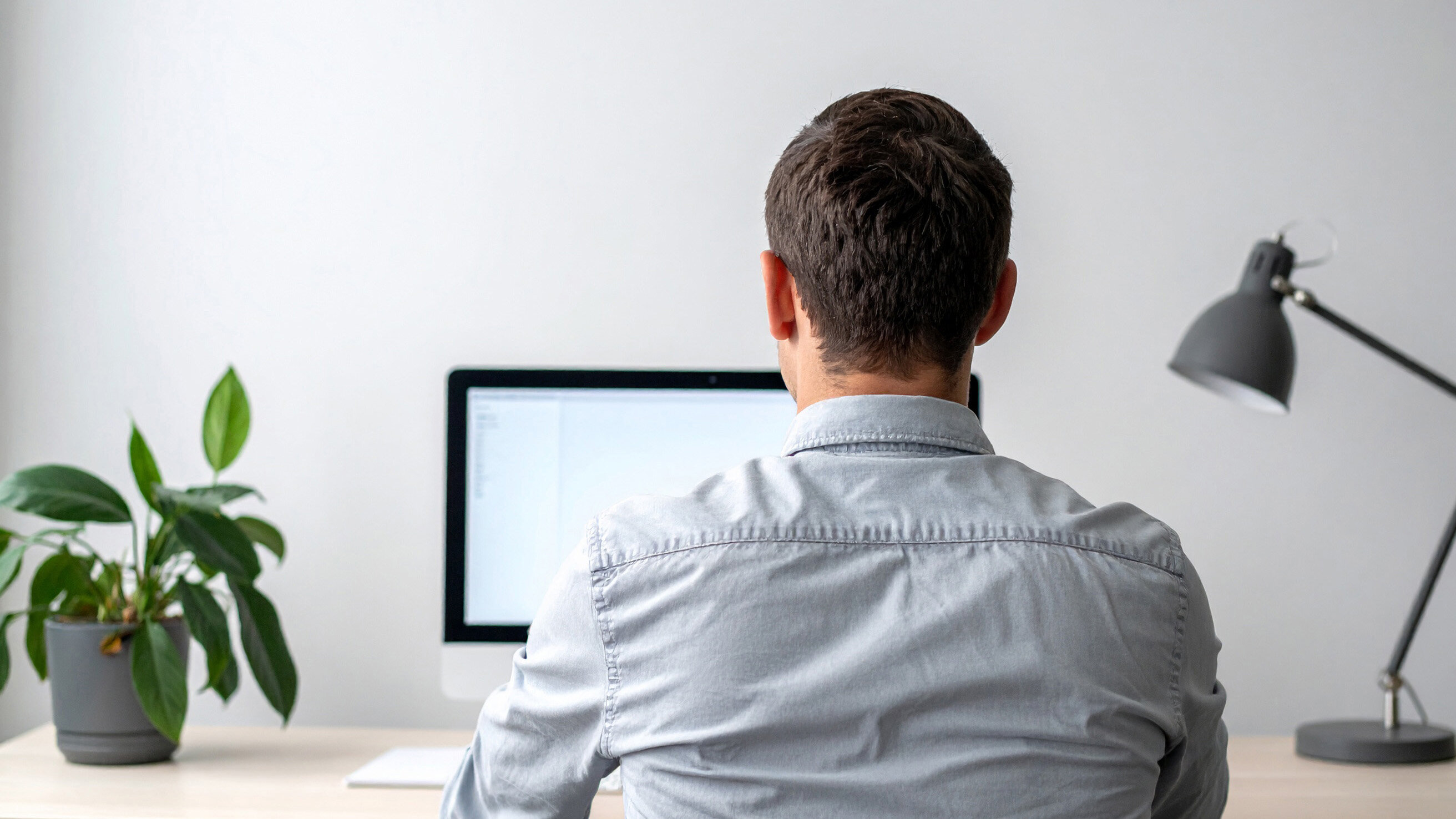 Ein Mann mit kurzen, braunen Haaren sitzt von hinten an einem Schreibtisch vor einem Computer. Auf dem Tisch steht eine grüne Pflanze und eine Schreibtischlampe.