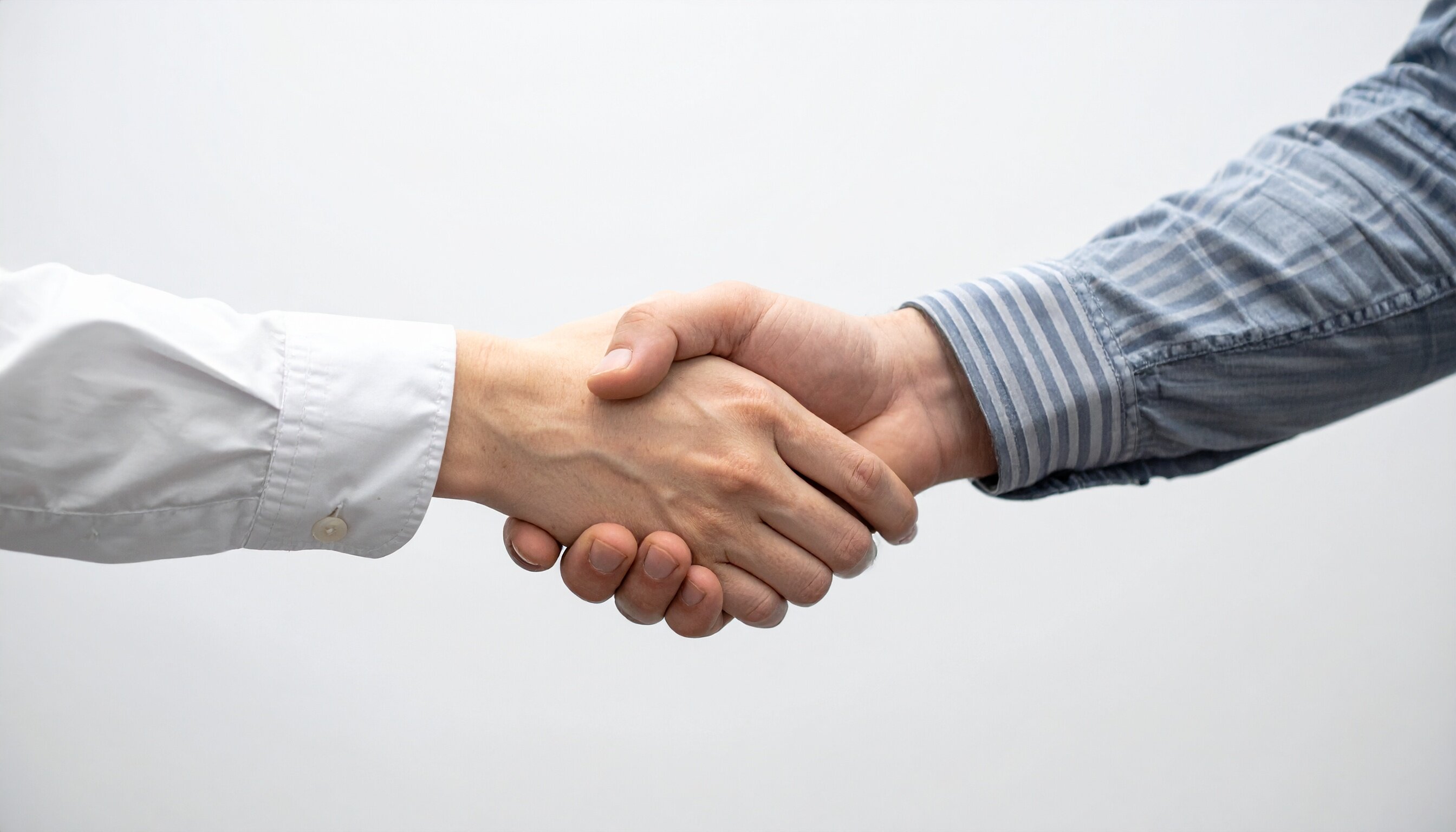 Zwei Hände schütteln sich, eine trägt ein weißes Hemd, die andere ein blau gestreiftes Hemd. Der Hintergrund ist neutral.