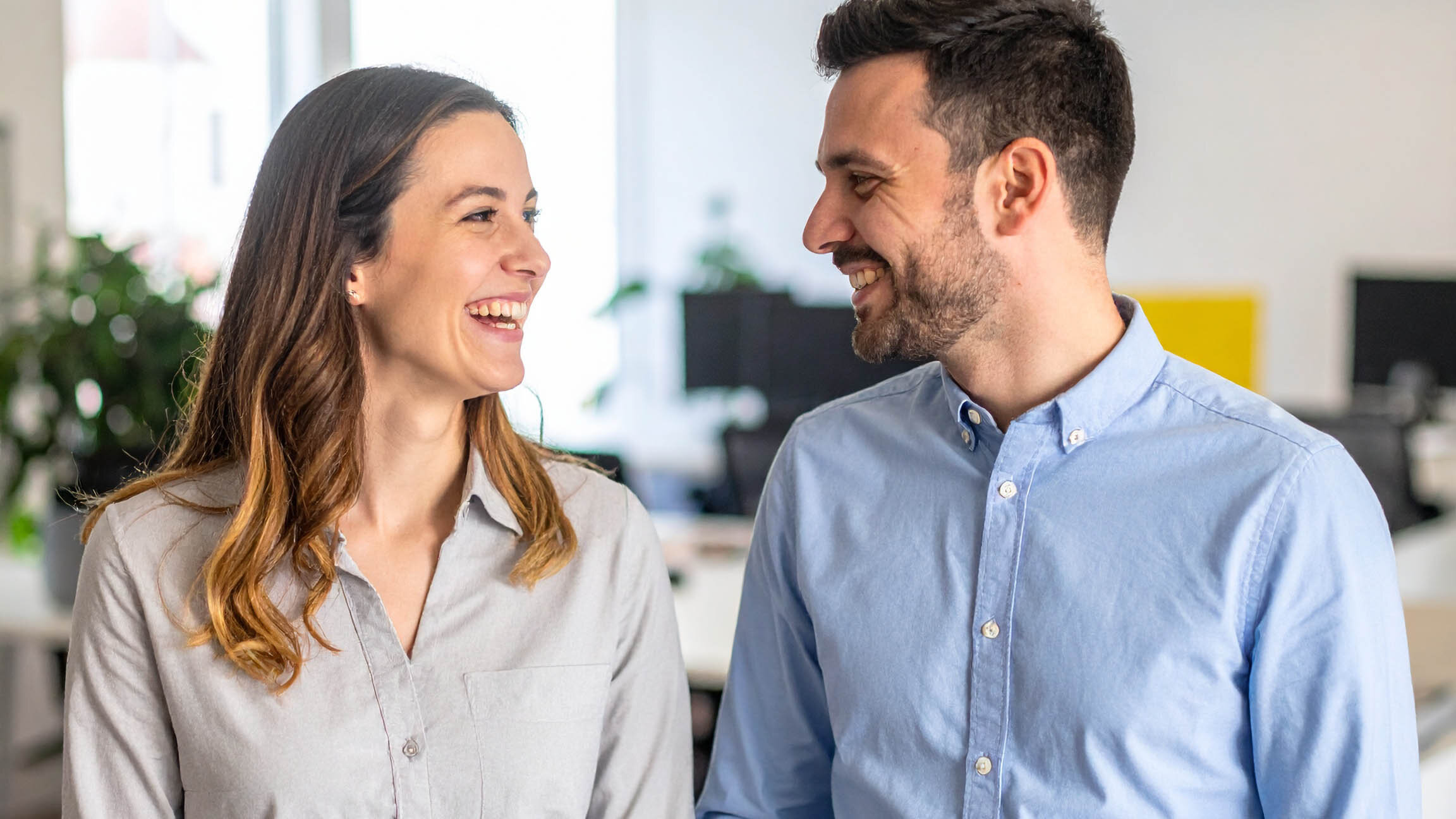 Zwei Personen stehen nebeneinander in einem modernen Büro. Die Person links trägt ein helles Hemd, die Person rechts ein blaues Hemd. Im Hintergrund sind Pflanzen und Büromöbel sichtbar.