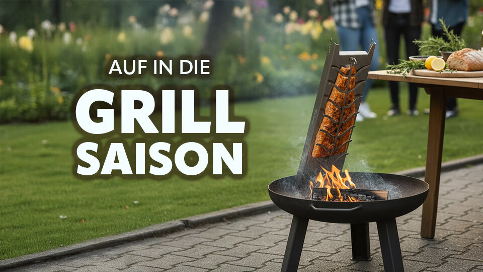 Mobiler Flammlachshalter mit brennendem Feuer und Lachsstücken vor einem Tisch im Garten mit Menschen im Hintergrund und dem Text 'AUF IN DIE GRILL SAISON'