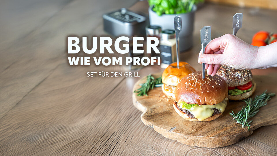 Drei Burger mit Käse und Sesambrötchen auf Holzbrett mit Kräutern und Hand, die einen Burger mit Metallspieß hält, Text 'BURGER WIE VOM PROFI SET FÜR DEN GRILL'