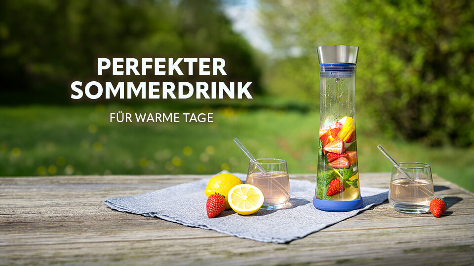 Glaskaraffe mit blauem Deckel und Boden gefüllt mit Wasser, Zitronenscheiben, Erdbeeren und Minzblättern auf einem grauen Tuch neben zwei Gläsern mit Strohhalmen, Zitronenhälfte und Erdbeere auf Holztisch im Freien