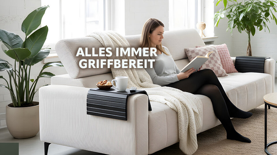 Frau sitzt auf hellgrauem Sofa mit Kissen und Decke liest Buch neben Pflanze und Beistelltisch mit Tasse und Snack