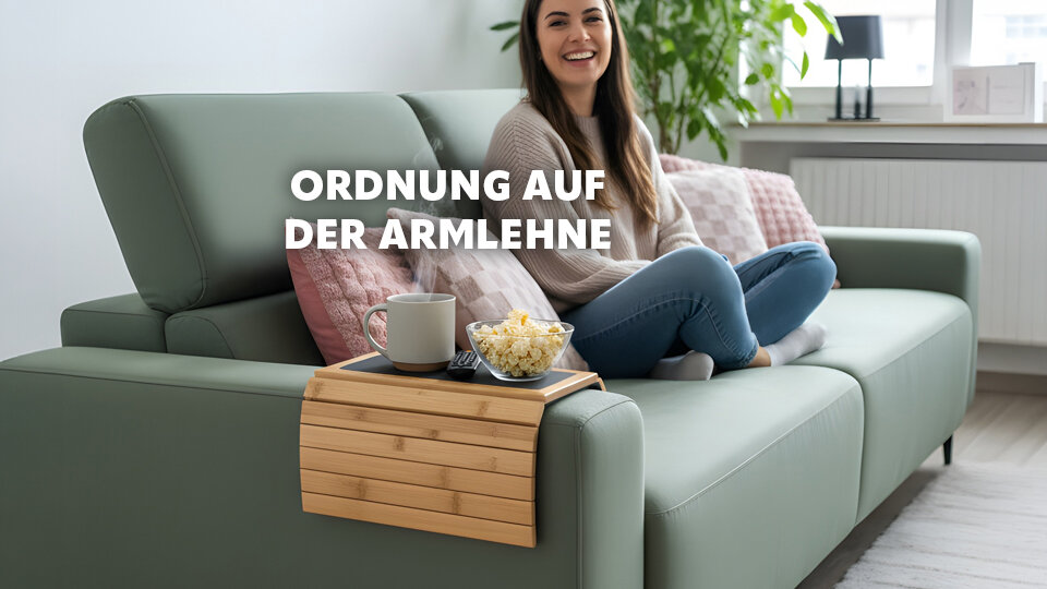 Frau sitzt lächelnd auf einem grünen Sofa mit einem Tablett auf der Armlehne, darauf eine Tasse und eine Schale Popcorn, im Hintergrund Fenster und Pflanze