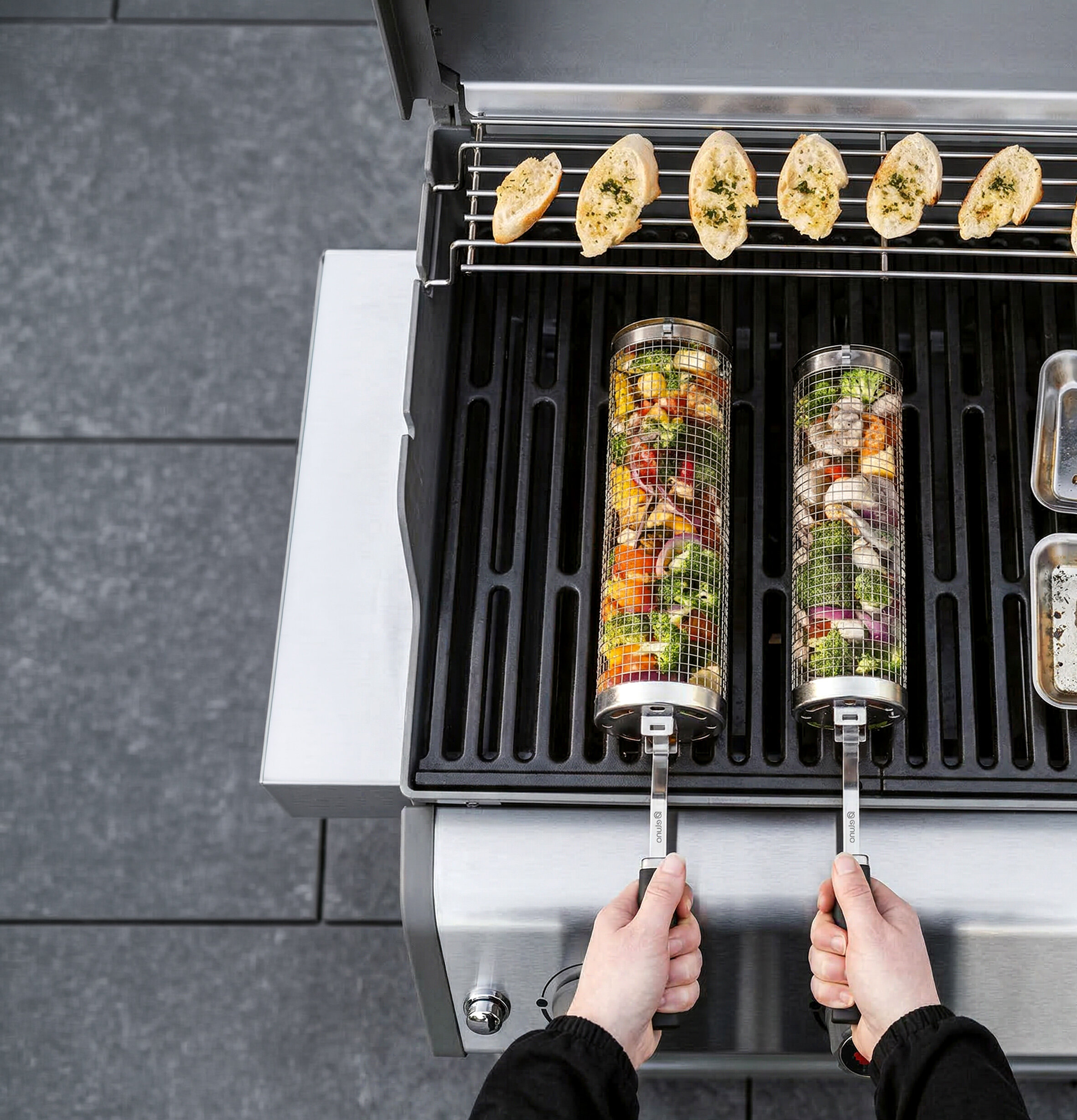 Zwei Grillkörbe mit Gemüse und Fleisch werden auf einem Gasgrill gehalten, darüber liegen sechs mit Kräutern belegte Baguettescheiben auf dem Grillrost