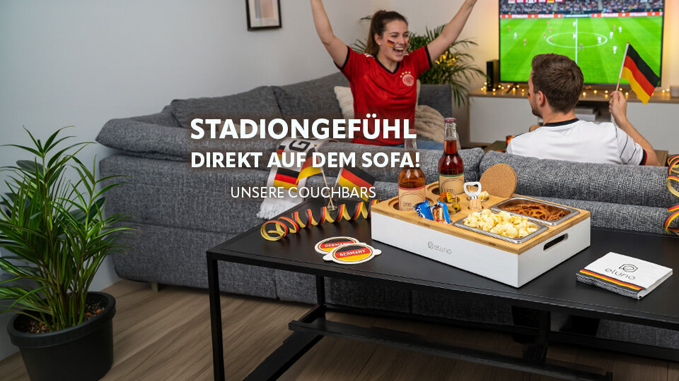 Gruppe von Menschen in Fußballtrikots jubelt vor einem Sofa mit Deutschlandflaggenkissen und einem Tisch mit Snacks und Getränken