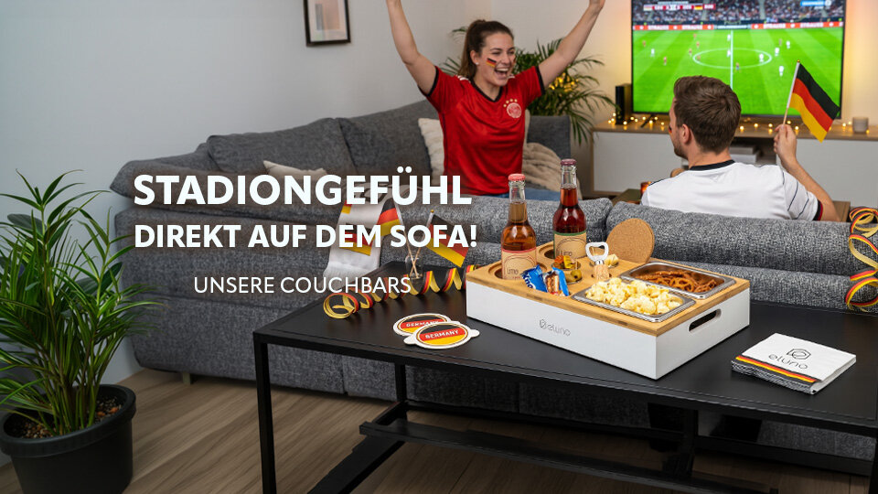 Gruppe von Menschen in Fußballtrikots jubelt vor einem Sofa mit Deutschlandflaggenkissen und einem Tisch mit Snacks und Getränken