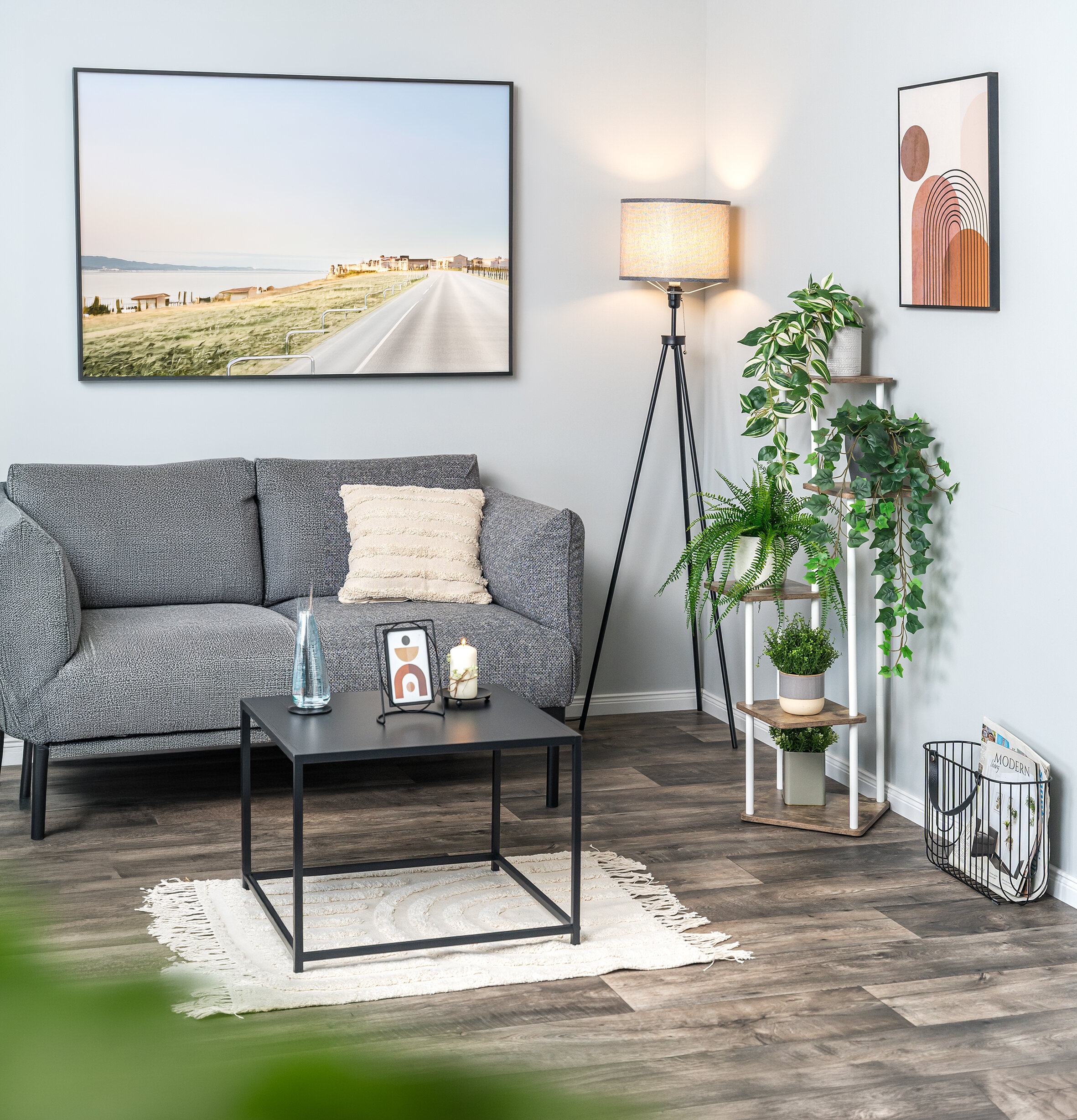 Wohnzimmer mit grauem Sofa, schwarzem quadratischem Couchtisch, Stehlampe, Pflanzenständer mit mehreren Pflanzen und abstrakten Wandbildern