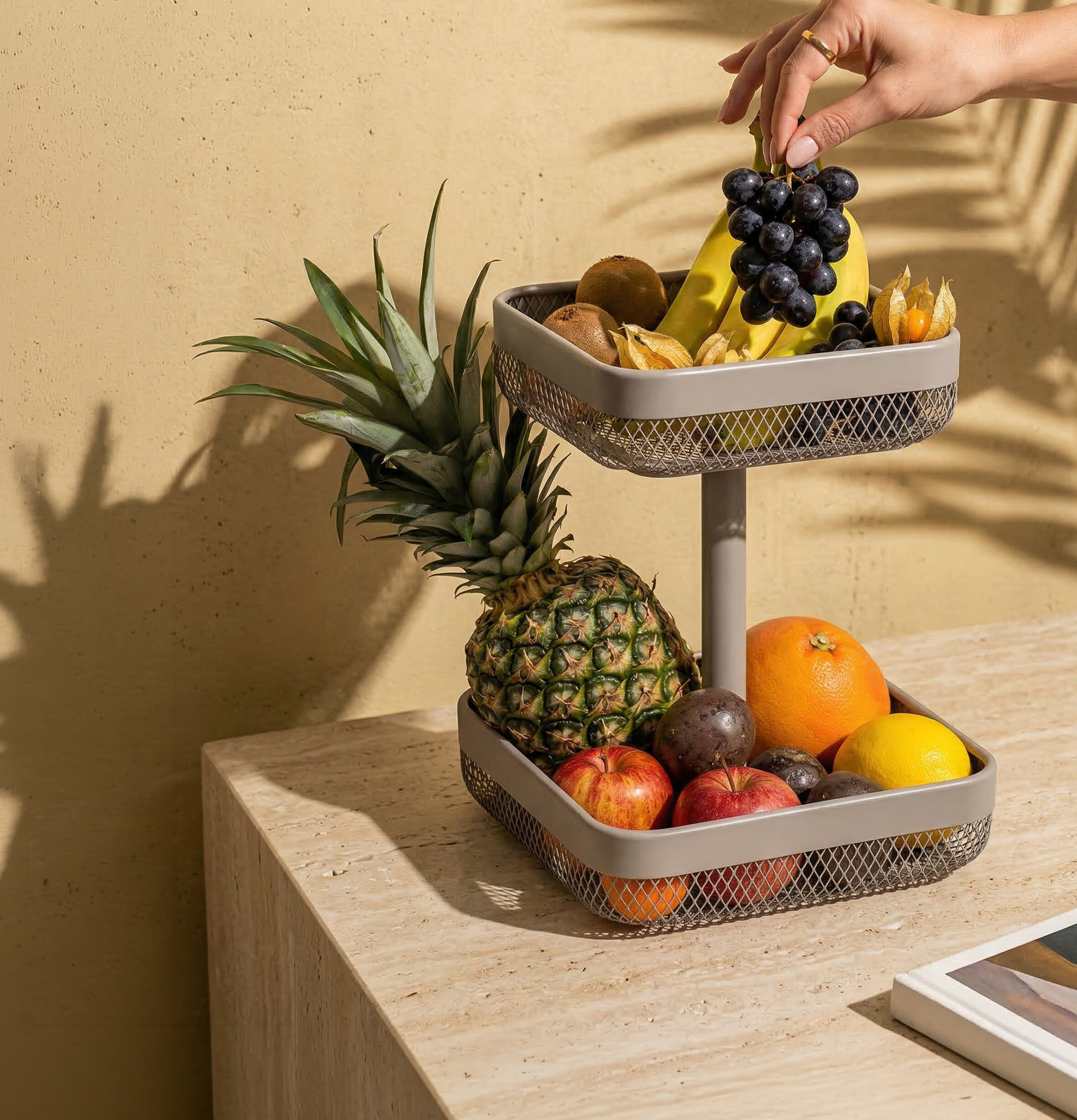 Zweistöckige Obstetagere mit Ananas, Äpfeln, Bananen, Trauben und weiteren Früchten auf einem hellen Tisch vor beiger Wand