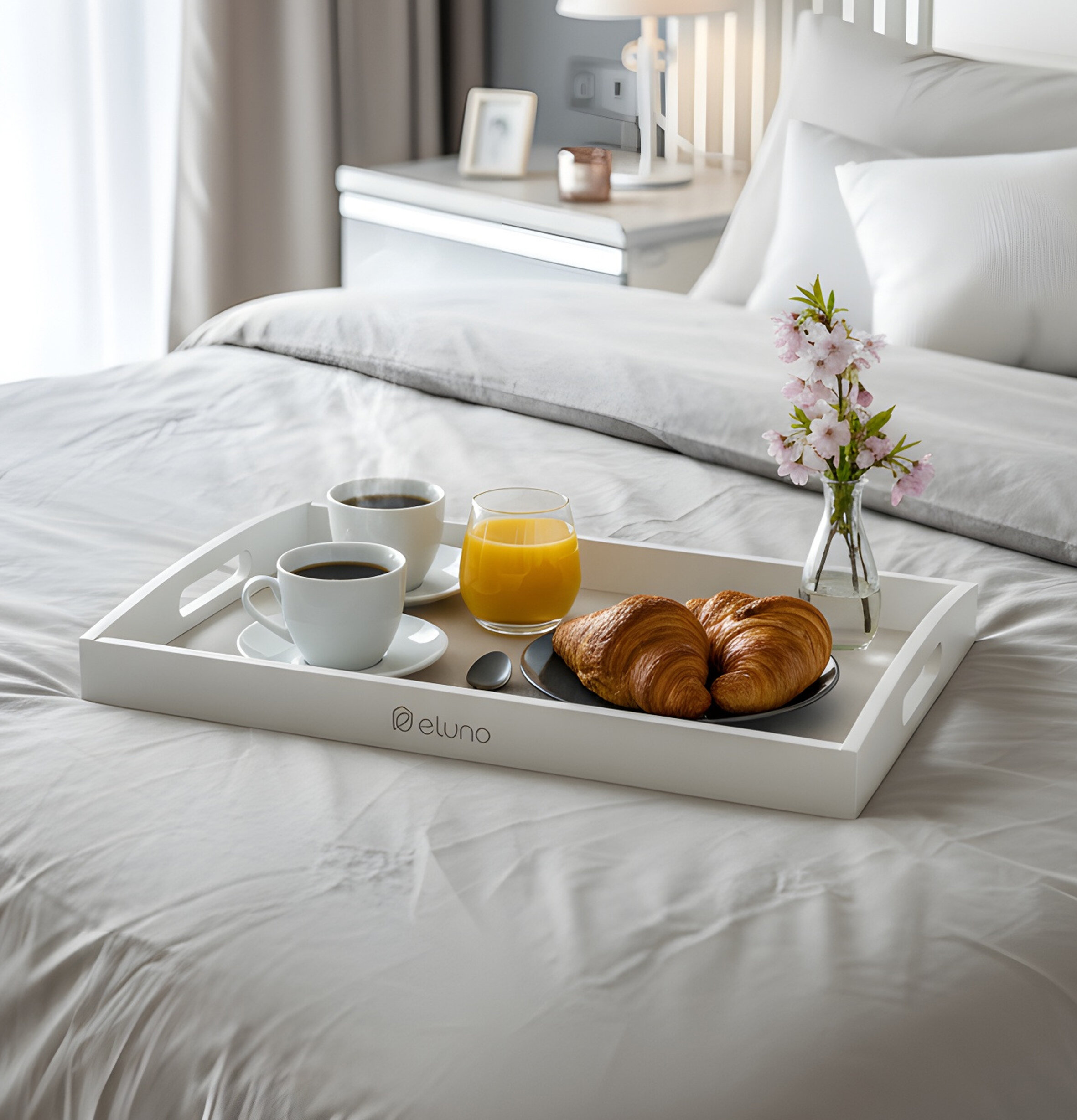 Weißes Serviertablett mit zwei Tassen Kaffee, Glas Orangensaft, zwei Croissants und Vase mit rosa Blüten auf Bett