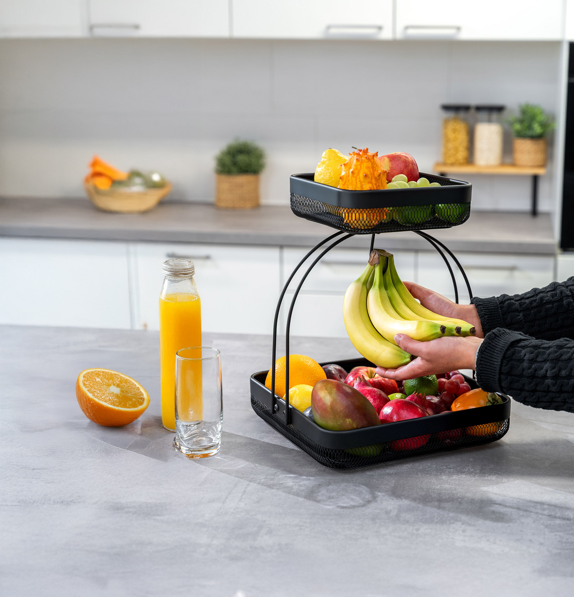 Zweistöckiger schwarzer Obstkorb mit verschiedenen Früchten auf einer Küchenarbeitsplatte, daneben eine Flasche Orangensaft, ein Glas und eine halbe Orange, Hände legen Bananen in den Korb